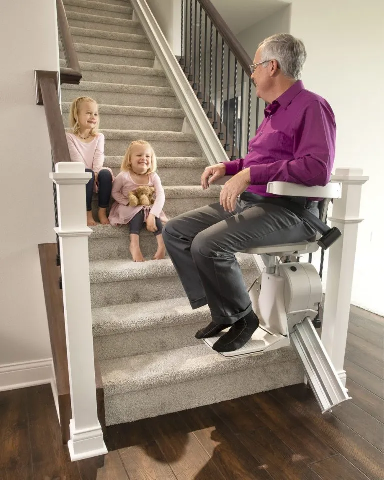 Senior man riding his Liftology stairlift while his two grandchildren wave from the carpeted stairs